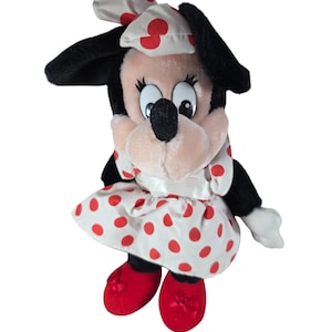Pu&ograve; includere: Una bambola di peluche di Minnie Mouse. La bambola ha orecchie nere, un viso rosa e un vestito bianco a pois rossi. Indossa un fiocco abbinato e scarpe rosse.