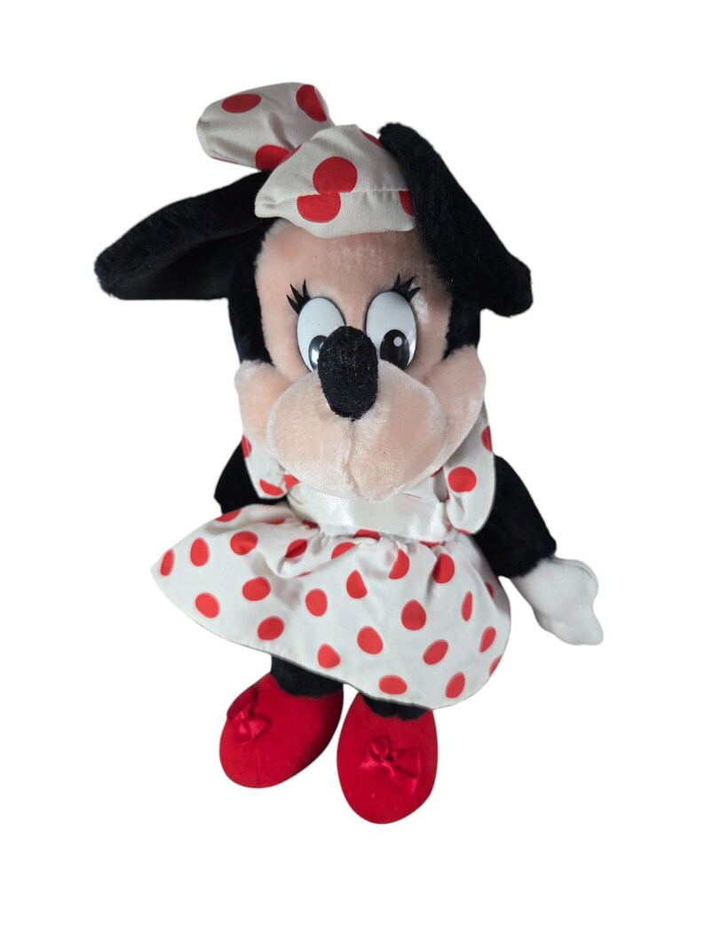 Pu&ograve; includere: Una bambola di peluche di Minnie Mouse. La bambola ha orecchie nere, un viso rosa e un vestito bianco a pois rossi. Indossa un fiocco abbinato e scarpe rosse.