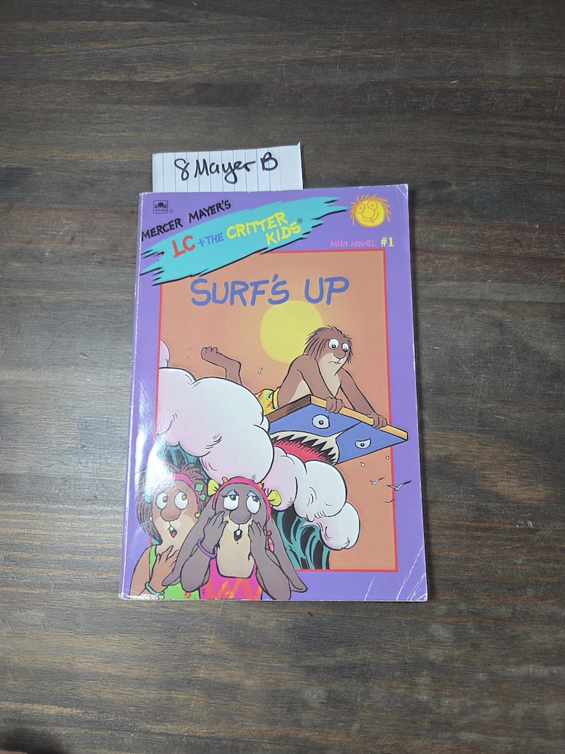 May include: A children's book titled "LC + The Critter Kids Surf's Up" by Mercer Mayer. The cover features cartoon characters surfing, with a purple border and a yellow sun. The book is a mini novel, number 1.
