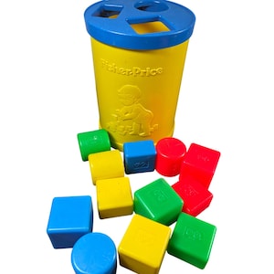 May include: A yellow Fisher-Price shape sorter toy with a blue lid and various colorful shapes. The shapes include red, green, yellow, and blue cubes, cylinders, and other geometric forms. The toy has the Fisher-Price logo.