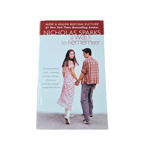 Peut inclure: Un livre de poche intitulé A Walk to Remember de Nicholas Sparks. La couverture montre un couple se tenant la main, la femme porte une robe à fleurs et l'homme une chemise à carreaux. Le texte inclut Now a Major Motion Picture et une citation.