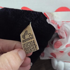 Pu&ograve; includere: Un peluche con corpo nero e piedi bianchi. Il giocattolo ha un motivo a pois bianchi e rossi sui suoi vestiti. &Egrave; visibile un'etichetta con la parola "applause".