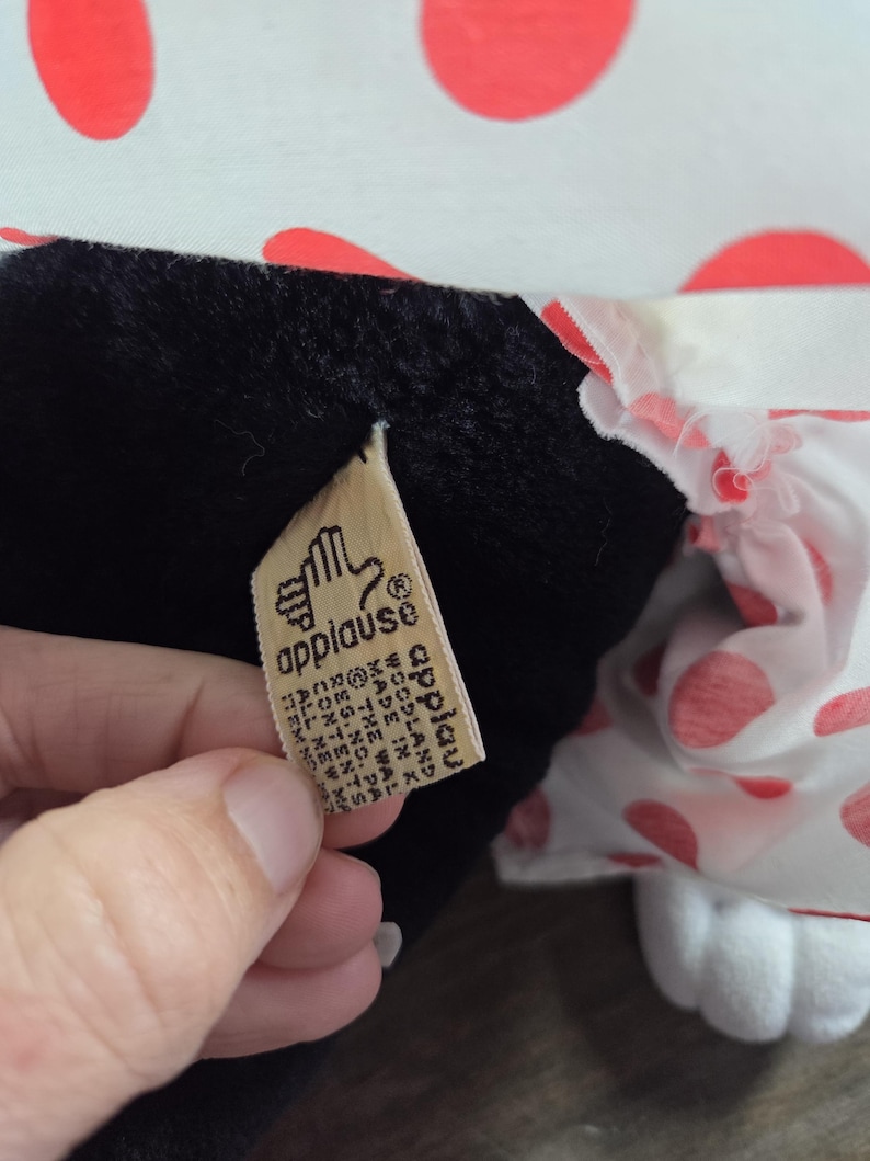 Pu&ograve; includere: Un peluche con corpo nero e piedi bianchi. Il giocattolo ha un motivo a pois bianchi e rossi sui suoi vestiti. &Egrave; visibile un'etichetta con la parola "applause".