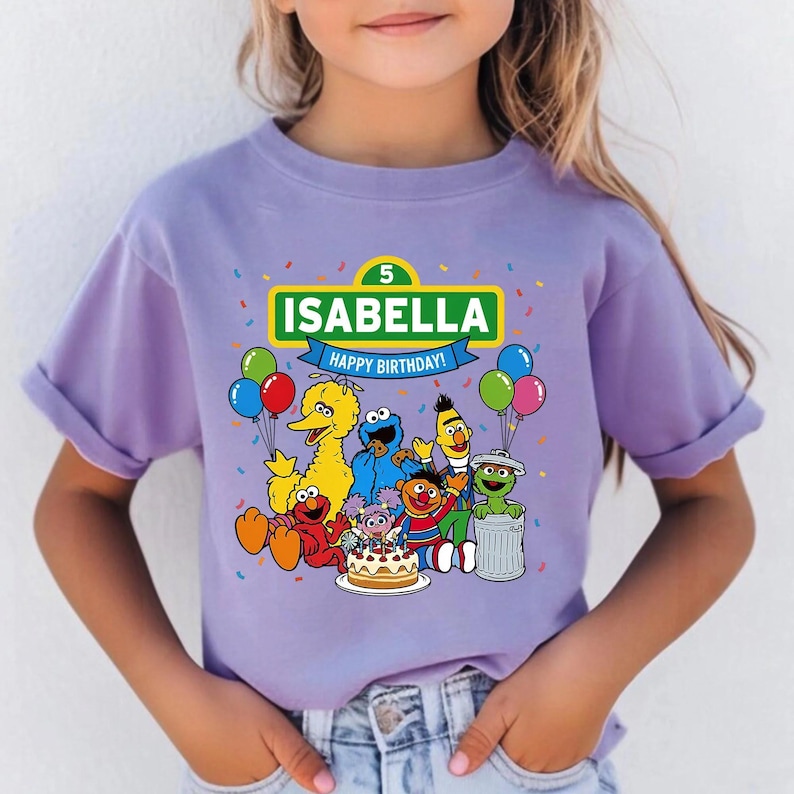 May include: A lilac-colored t-shirt featuring the name "ISABELLA" above a "Happy Birthday!" banner, with the number "5". The shirt showcases Sesame Street characters, balloons, and confetti, creating a festive birthday theme.