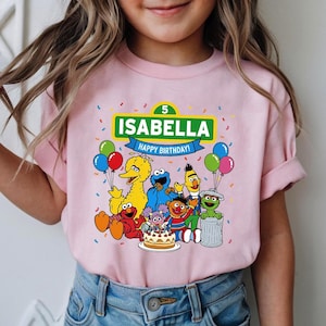 May include: Pink t-shirt featuring a colorful graphic of Sesame Street characters, a birthday cake, and balloons. The shirt has the name "Isabella" and the words "Happy Birthday!" printed on it. The number "5" is also displayed.