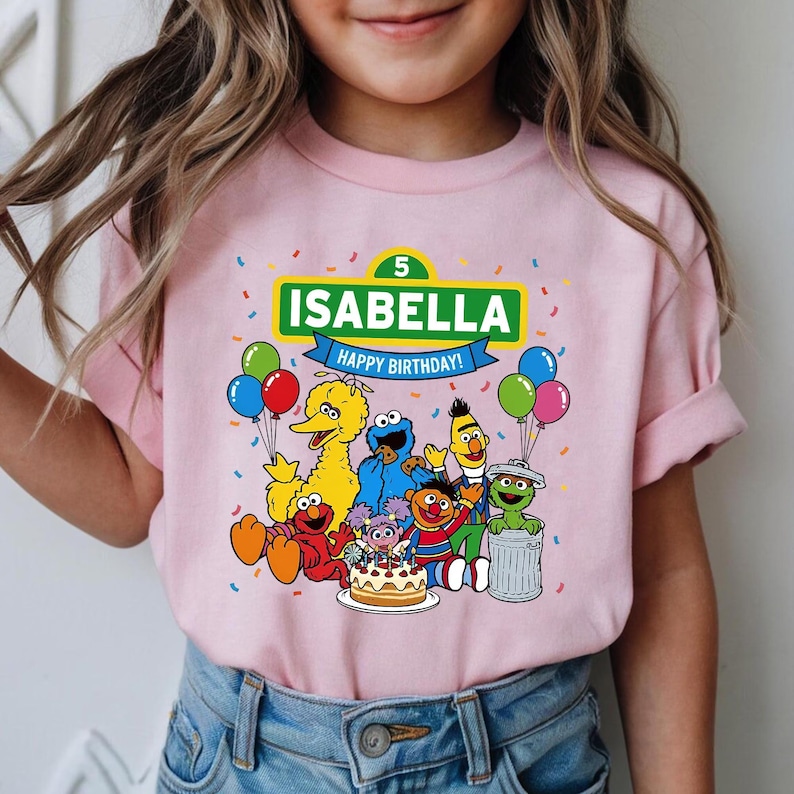 May include: Pink t-shirt featuring a colorful graphic of Sesame Street characters, a birthday cake, and balloons. The shirt has the name "Isabella" and the words "Happy Birthday!" printed on it. The number "5" is also displayed.