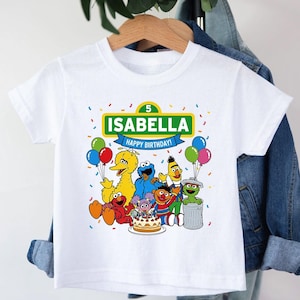 May include: White t-shirt featuring a colorful graphic of Sesame Street characters, balloons, and a birthday cake. The shirt has the name "ISABELLA" and "HAPPY BIRTHDAY!" printed on it, with the number "5" above the name.