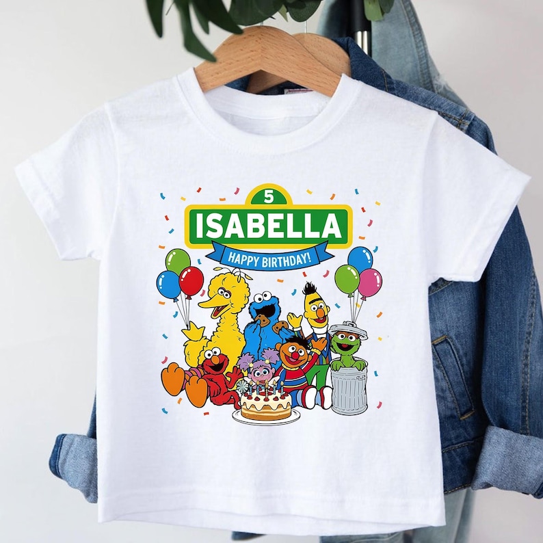 May include: White t-shirt featuring a colorful graphic of Sesame Street characters, balloons, and a birthday cake. The shirt has the name "ISABELLA" and "HAPPY BIRTHDAY!" printed on it, with the number "5" above the name.