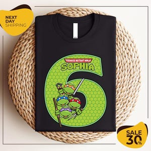 May include: Black t-shirt featuring a green number six design with the Teenage Mutant Ninja Turtles. The design includes the name "Sophia" and the turtles holding weapons. The shirt is displayed on a woven surface.