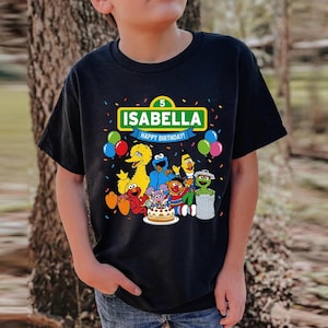 May include: Black t-shirt featuring a colorful graphic of Sesame Street characters, a birthday cake, and the name "ISABELLA" with "HAPPY BIRTHDAY!" text. The number "5" is also displayed. Balloons and confetti surround the design.