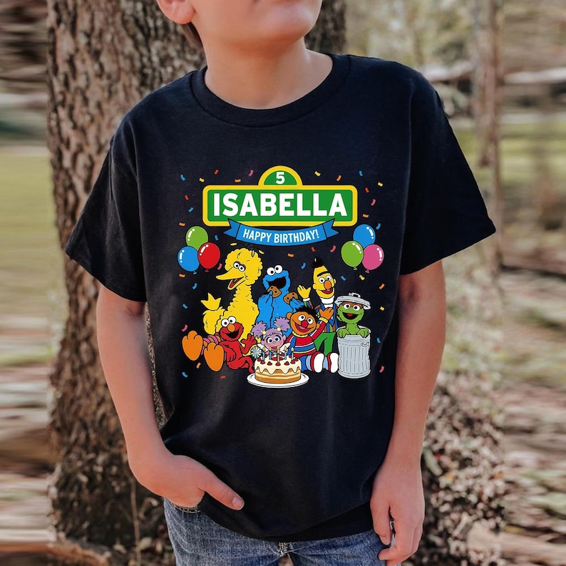 May include: Black t-shirt featuring a colorful graphic of Sesame Street characters, a birthday cake, and the name "ISABELLA" with "HAPPY BIRTHDAY!" text. The number "5" is also displayed. Balloons and confetti surround the design.
