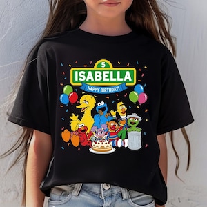 May include: Black t-shirt featuring a colorful graphic of Sesame Street characters, a birthday cake, and balloons. The shirt has the name "Isabella" and the text "Happy Birthday!" printed on it.