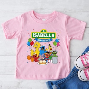May include: Pink short-sleeved t-shirt featuring a graphic of Sesame Street characters. The shirt includes the name "ISABELLA" and the text "HAPPY BIRTHDAY!" above the characters. Colorful balloons and confetti are also present in the design.