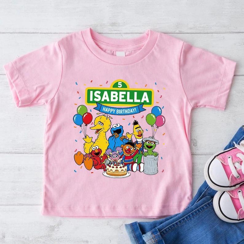 May include: Pink short-sleeved t-shirt featuring a graphic of Sesame Street characters. The shirt includes the name "ISABELLA" and the text "HAPPY BIRTHDAY!" above the characters. Colorful balloons and confetti are also present in the design.
