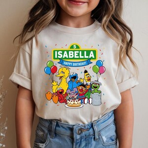 May include: Beige t-shirt featuring a colorful Sesame Street graphic. The design includes the name "Isabella" and the text "Happy Birthday!" with the number 5. The shirt showcases various Sesame Street characters, balloons, and confetti.