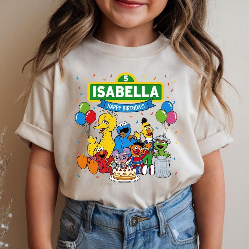 May include: Beige t-shirt featuring a colorful Sesame Street graphic. The design includes the name "Isabella" and the text "Happy Birthday!" with the number 5. The shirt showcases various Sesame Street characters, balloons, and confetti.