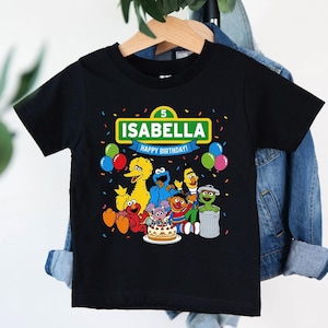 May include: Black t-shirt featuring a colorful graphic of Sesame Street characters. The design includes the name "ISABELLA" above "HAPPY BIRTHDAY!" with a number 5. Balloons and confetti add to the festive birthday theme.