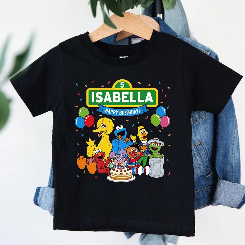 May include: Black t-shirt featuring a colorful graphic of Sesame Street characters. The design includes the name "ISABELLA" above "HAPPY BIRTHDAY!" with a number 5. Balloons and confetti add to the festive birthday theme.
