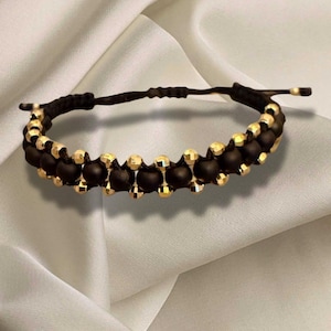 Könnte beinhalten: Ein Armband mit schwarzen und goldenen Perlen. Das Armband hat abwechselnde Reihen aus schwarzen und goldenen Perlen. Die goldenen Perlen haben ein strukturiertes, metallisches Aussehen. Das Armband ist vor einem weißen, seidigen Hintergrund platziert.