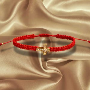 Könnte beinhalten: Ein rotes Makramee-Armband mit einem goldfarbenen Kreuzanhänger. Das Armband wird auf einer Satin-Oberfläche präsentiert. Das Kreuz ist mit kleinen, funkelnden Verzierungen versehen.