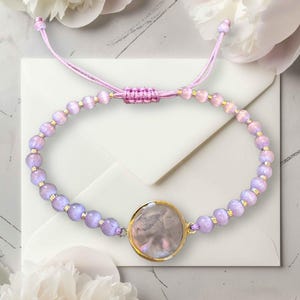 Könnte beinhalten: Ein Armband mit einem zentralen, runden Anhänger in Perlmuttoptik in einer goldfarbenen Fassung, flankiert von hellvioletten Perlen und kleinen goldfarbenen Zwischenelementen. Das Armband hat einen hellvioletten Kordelverschluss.