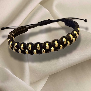 May include: Black and gold beaded bracelet. The bracelet features a series of black beads interspersed with gold-colored beads. The adjustable closure is black. The bracelet is displayed on a white, textured surface.