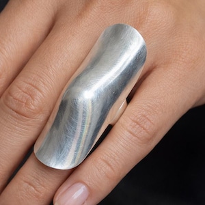Puede incluir: Un anillo plateado grande con un diseño curvo. El anillo es liso y pulido, reflejando la luz. El anillo cubre una parte significativa del dedo, mostrando un estilo de joyería moderno y audaz.