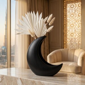 May include: A matte black ceramic vase shaped like a crescent moon, holding a collection of dried white palm spears and fluffy bunny tail grass stems. The vase is positioned on a polished marble surface with a smooth, curved silhouette.