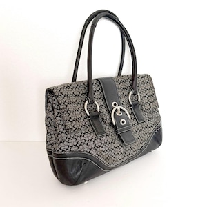 May include: A black and gray handbag featuring a repeating monogram pattern on the fabric body. The bag includes black leather trim, two top handles, and a central vertical leather strap secured with a silver-toned metal buckle hardware.
