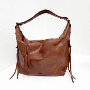May include: A brown leather hobo bag with a single shoulder strap. The bag has a slouchy design with long, thin leather tassels hanging from the sides. The strap is a darker brown with a textured pattern.