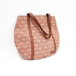 May include: A woven tote bag with a geometric pattern in red and beige. The bag features brown leather handles and trim. The tote is designed with a spacious interior and is suitable for carrying various items.