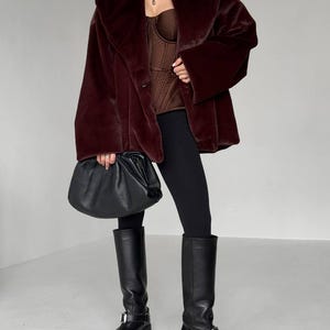 May include: A burgundy faux fur coat with a wide collar and long sleeves. The outfit includes a brown corset top, black leggings, and knee-high black leather boots. A black leather handbag is held in the hand.