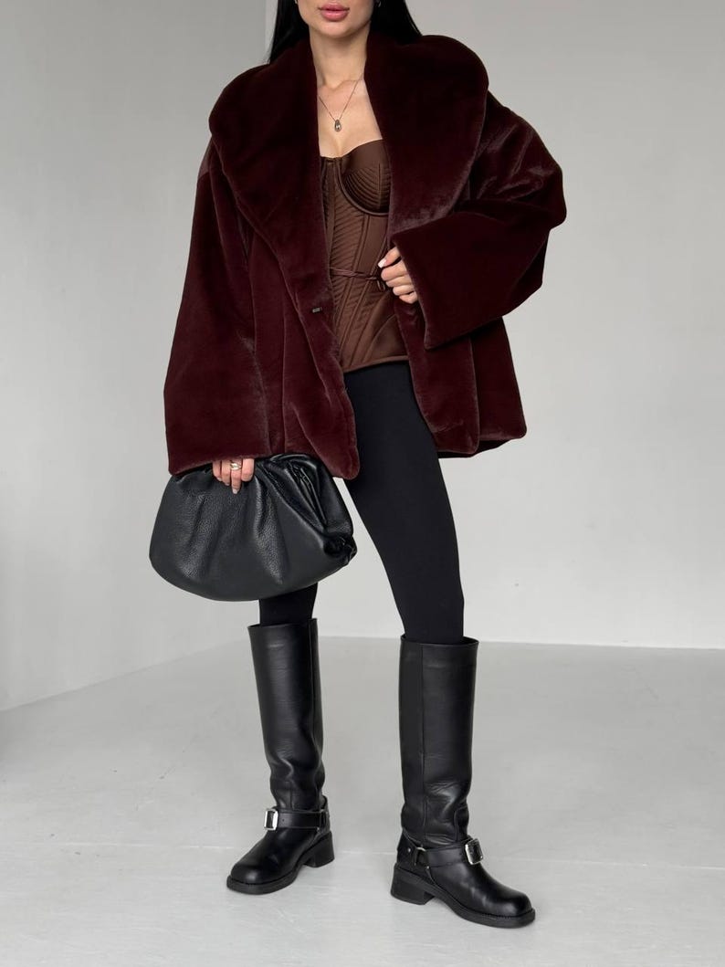 May include: A burgundy faux fur coat with a wide collar and long sleeves. The outfit includes a brown corset top, black leggings, and knee-high black leather boots. A black leather handbag is held in the hand.