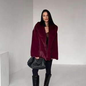 May include: A burgundy faux fur coat with a wide collar. The coat is paired with black leggings and knee-high black leather boots. A black, gathered leather handbag is held in the hand.