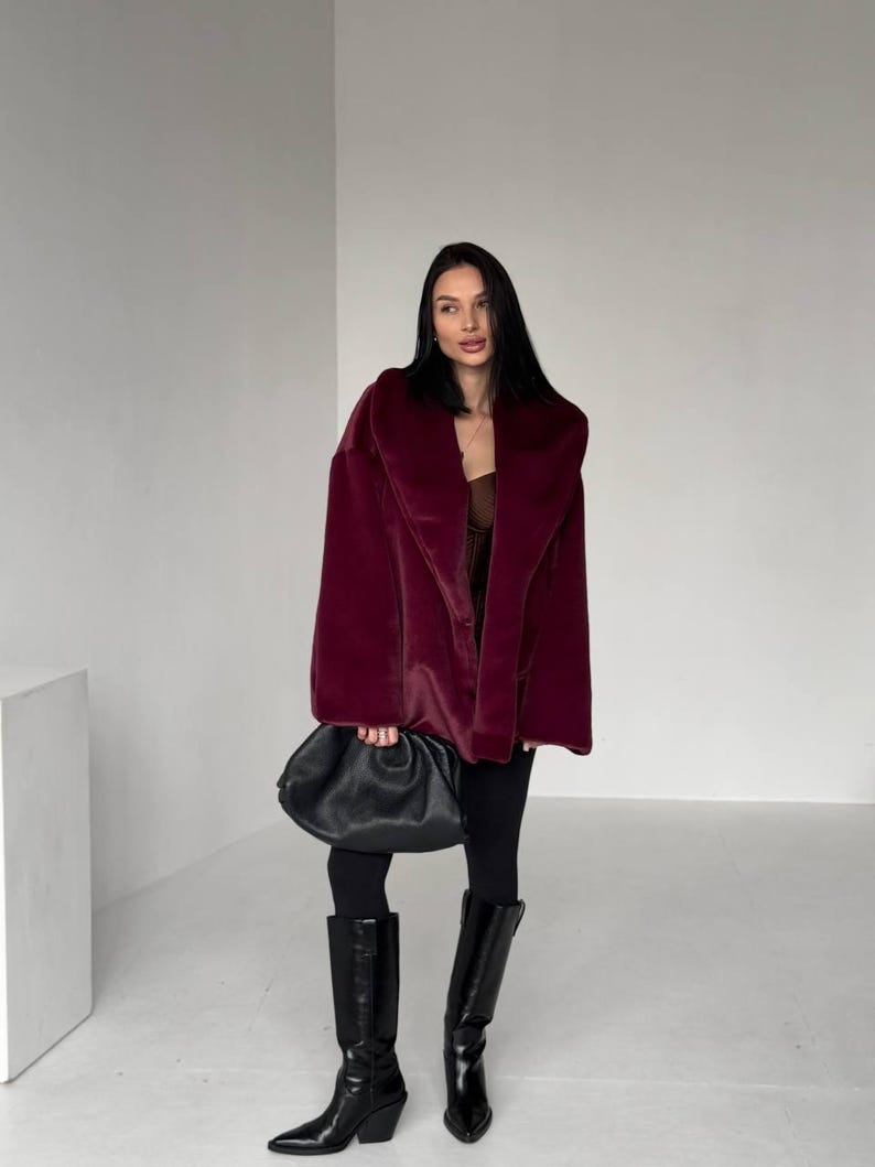 May include: A burgundy faux fur coat with a wide collar. The coat is paired with black leggings and knee-high black leather boots. A black, gathered leather handbag is held in the hand.