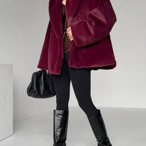 May include: A burgundy faux fur coat with a wide collar and long sleeves. The coat is paired with black leggings and knee-high black leather boots. A black handbag is held in the left hand. The outfit is set against a white background.