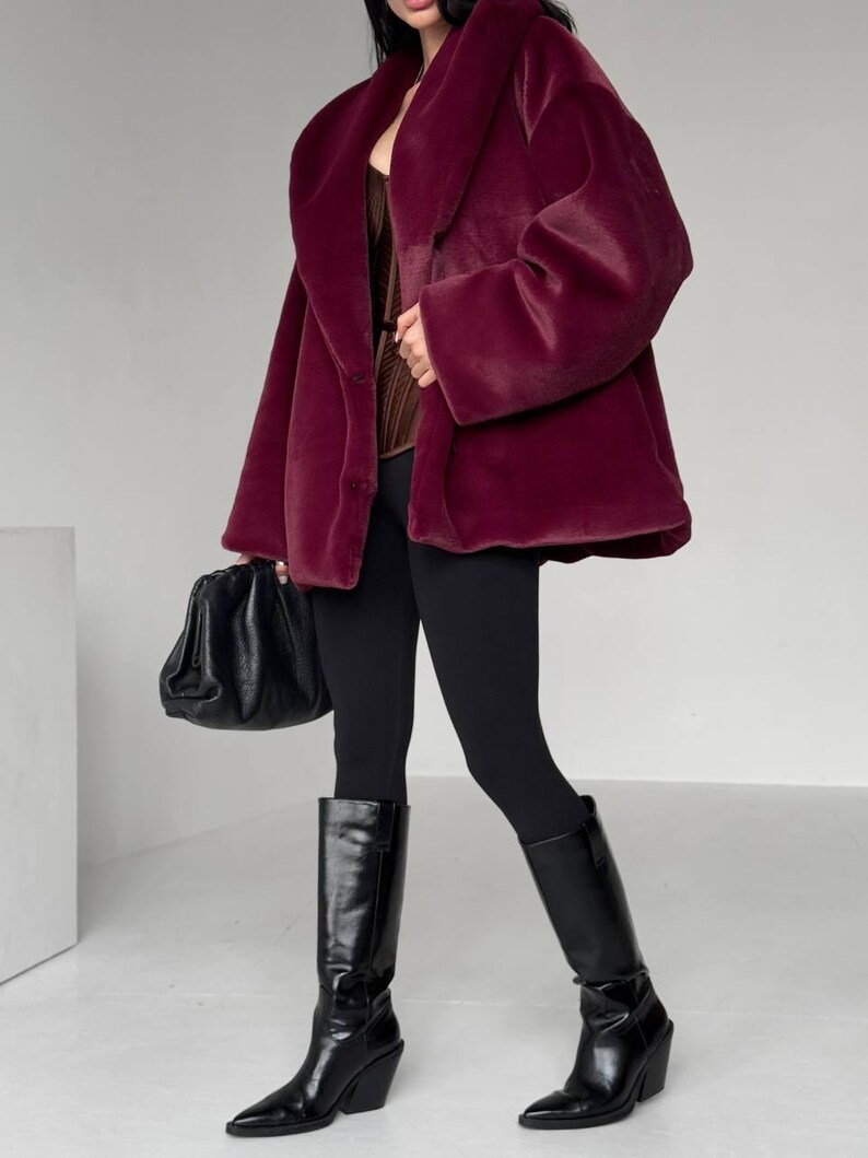 May include: A burgundy faux fur coat with a wide collar and long sleeves. The coat is paired with black leggings and knee-high black leather boots. A black handbag is held in the left hand. The outfit is set against a white background.