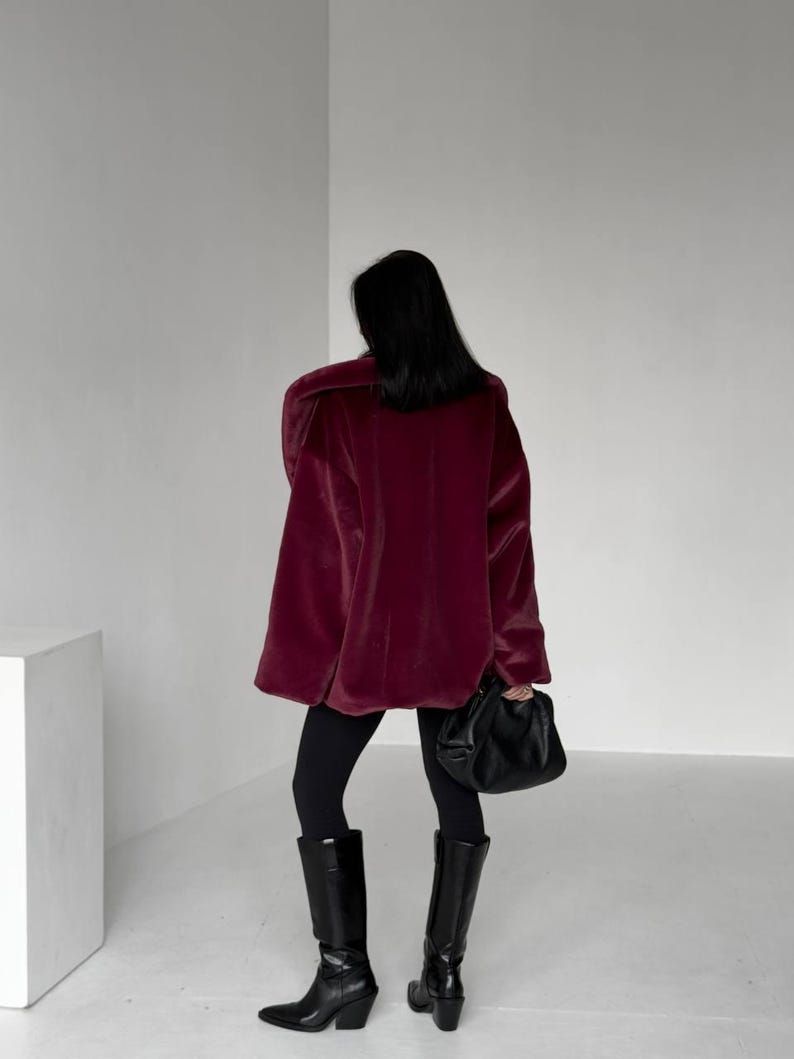 May include: A person wearing a burgundy oversized coat, black leggings, and black knee-high boots. The person is holding a black handbag. The coat has a plush, velvety texture. The boots have a pointed toe and a block heel.