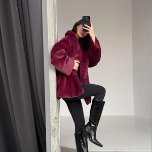 May include: A burgundy faux fur coat with a hood. The coat is paired with black leggings and black knee-high boots. The boots have a pointed toe and a block heel. The background is a neutral gray and white.