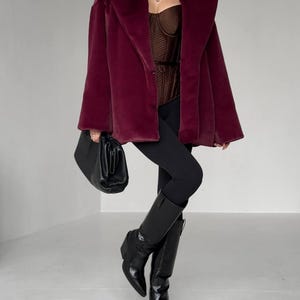 May include: A burgundy faux fur coat with a wide collar. The coat is paired with a brown corset, black leggings, and black knee-high boots. A black handbag is held in the left hand. The outfit is set against a white background.