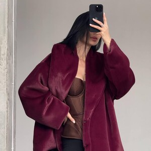 May include: A burgundy faux fur coat with a wide collar and long sleeves. The coat is buttoned and worn over a brown corset top and black pants. The person is holding a black phone in front of their face.