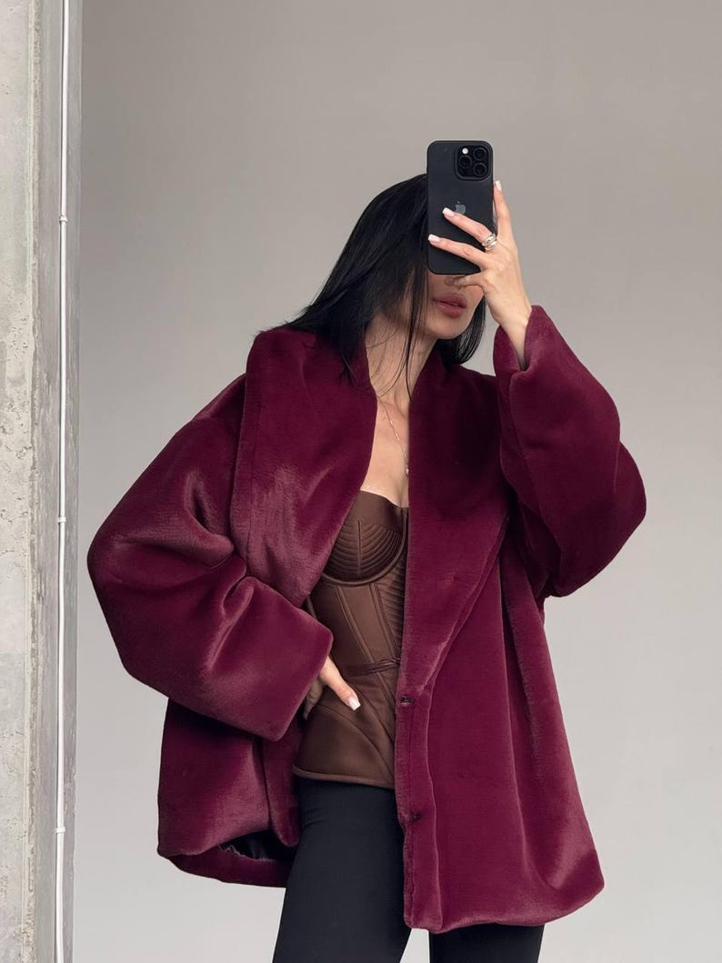 May include: A burgundy faux fur coat with a wide collar and long sleeves. The coat is buttoned and worn over a brown corset top and black pants. The person is holding a black phone in front of their face.