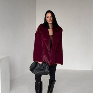 May include: A burgundy faux fur coat with a wide collar. The coat is paired with black leggings and knee-high black leather boots. A black, gathered leather handbag is held in the hand.