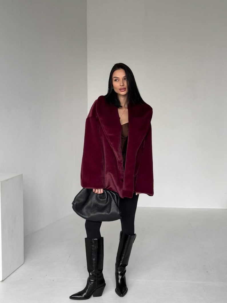 May include: A burgundy faux fur coat with a wide collar. The coat is paired with black leggings and knee-high black leather boots. A black, gathered leather handbag is held in the hand.