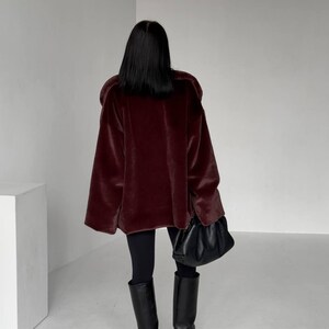 May include: A burgundy faux fur coat with wide sleeves. The coat is paired with black leggings and knee-high black leather boots. A black leather handbag is held in the right hand.