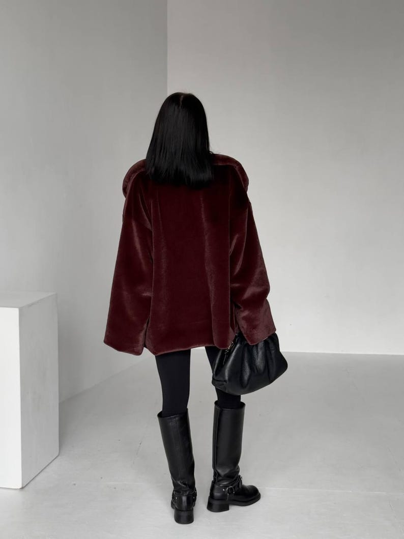 May include: A burgundy faux fur coat with wide sleeves. The coat is paired with black leggings and knee-high black leather boots. A black leather handbag is held in the right hand.