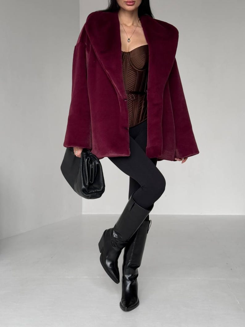 May include: A burgundy faux fur coat with a wide collar. The coat is paired with a brown corset, black leggings, and black knee-high boots. A black handbag is held in the left hand. The outfit is set against a white background.
