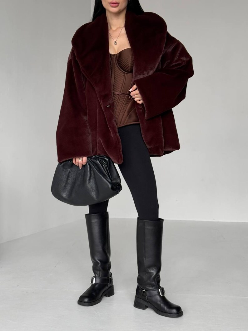 May include: A burgundy faux fur coat with a wide collar and long sleeves. The outfit includes a brown corset top, black leggings, and knee-high black leather boots. A black leather handbag is held in the hand.