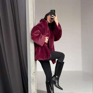May include: A burgundy faux fur coat with a hood. The coat is paired with black leggings and black knee-high boots. The boots have a pointed toe and a block heel. The background is a neutral gray and white.