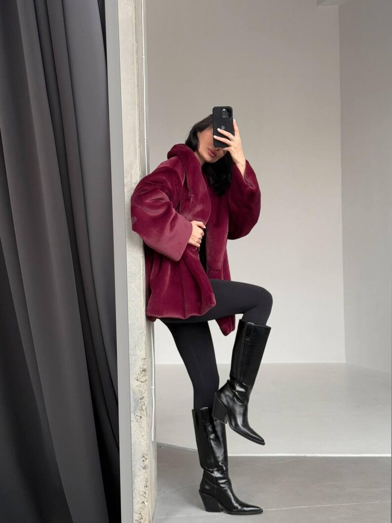 May include: A burgundy faux fur coat with a hood. The coat is paired with black leggings and black knee-high boots. The boots have a pointed toe and a block heel. The background is a neutral gray and white.
